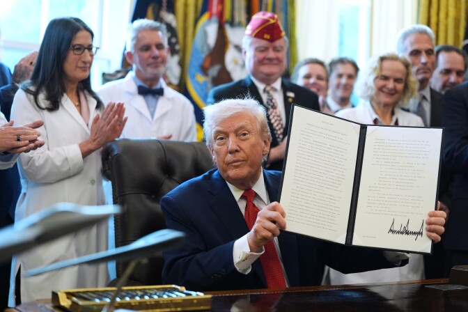 An older white man in a suit holds up a signed document as a crowd around him claps.