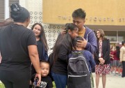 An adult with medium skin tone embraces two children outside a school. Adults and children mix nearby and in the background.