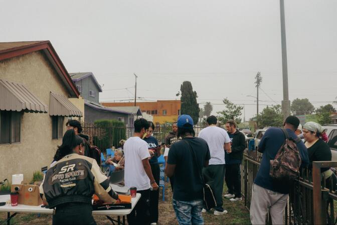 A group of people stand on the lawn of a home chatting. There's a table and a group of blue plastic chairs set up on the lawn
