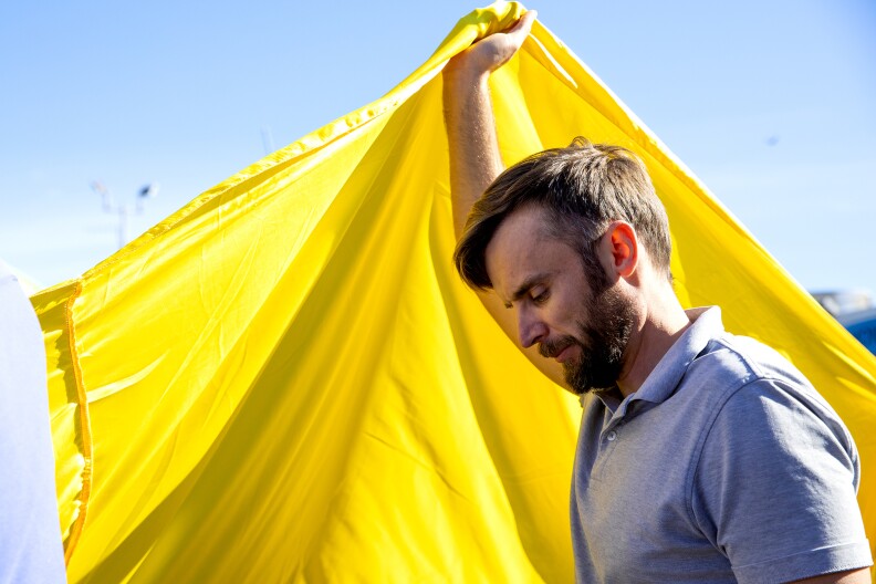A Caucasian man with medium-length brunette hair, a beard, and a gray polo shirt looks down with his eyes mostly closed outside in the daylight while holding up a yellow flag.