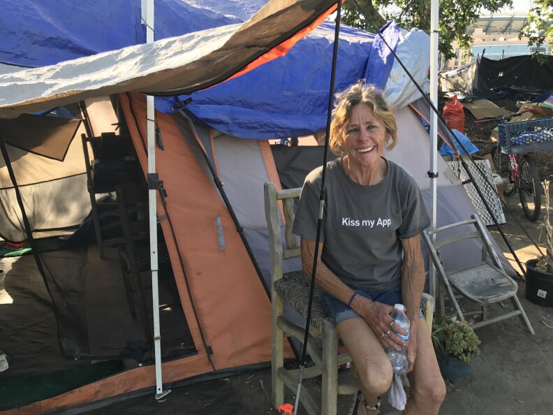 A light-skinned woman with blond hair in a ponytail and a gray shirt that says "Kiss my App" sits on a wooden chair in front of a tent. She's smiling into the camera.