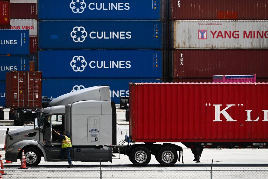 A truck driver wearing a yellow and orange safety vest climbs into a gray or silver colored cab of the truck. The truck's back has a red colored container with the letter 'K' on it. Behind the truck in the background are other shipping containers