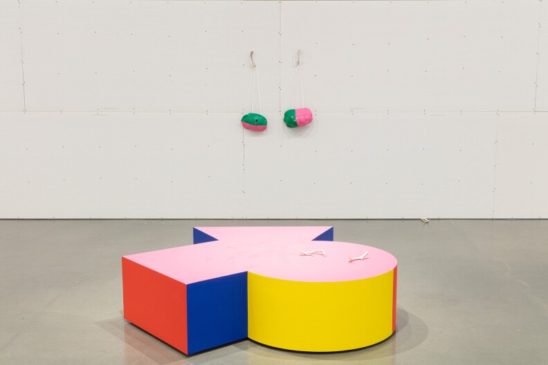 Plataforma con forma geométrica irregular, tapa rosa con laterales en rojo, azul y amarillo. En la parte circular de la plataforma hay un par de astas de ciervo. Colgados en la pared sin terminar en el fondo hay un par de caparazones de tortuga pintados de verde y rosa, colgados de cuerdas de la pared por astas de venado.