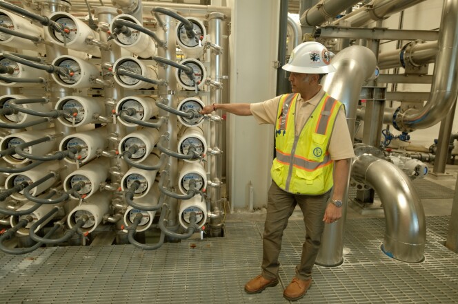 A man in a white hardhat and neon safety vest points to a machine that puts wastewater through reverse osmosis to purify it. 