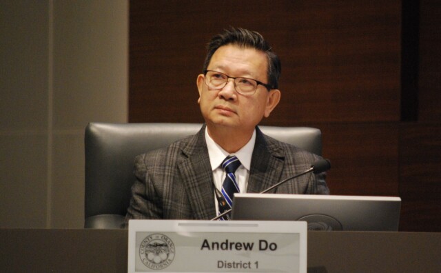 A man dressed in a suit jacket and tie looks up while seated in front of a sign that says "County of Orange California," "Andrew Do," "District 1."