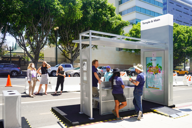 Three people stand under a bus shelter on a sidewalk, as three other people stand in the background.