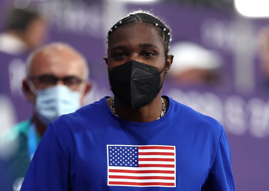 A man has a black mask on his face and is wearing a blue shirt with an American flag in the middle of it