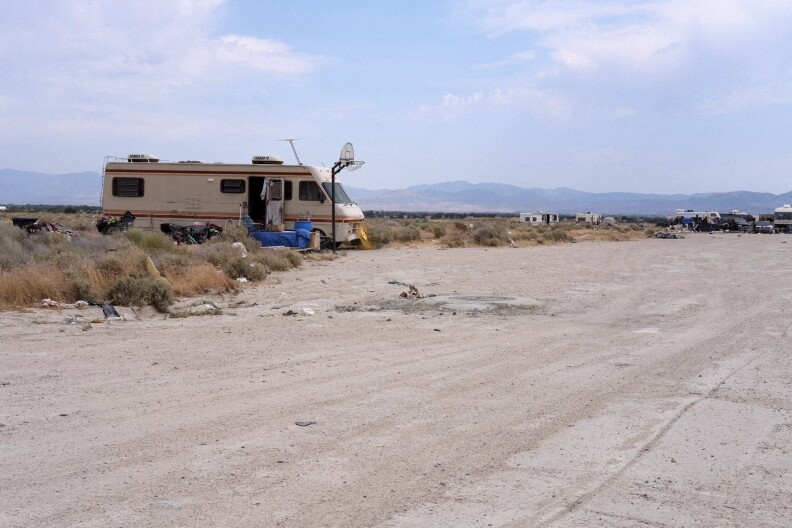 A camper is parked next to a basketball hoop in the middle of the desert. More campers are visible in the distance.