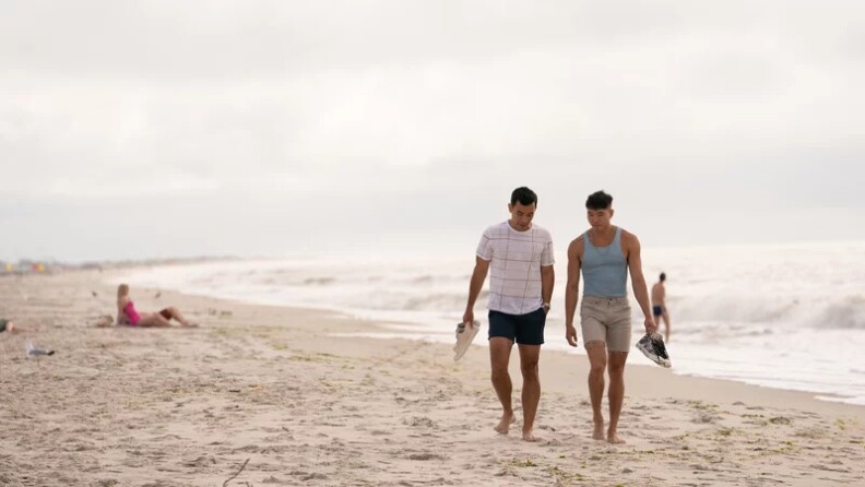 Two men who appear to be Asian American Pacific Islander and in their 20s or 30s walk closely together on a beach. They each hold their shoes. The day is overcast.