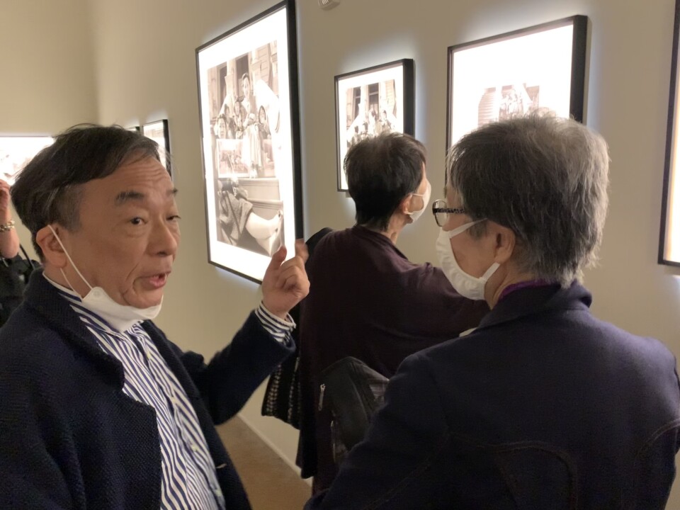 A Japanese man in his 60's points to black-and-white photos on the walls of a musuem, while a visitor watches him.