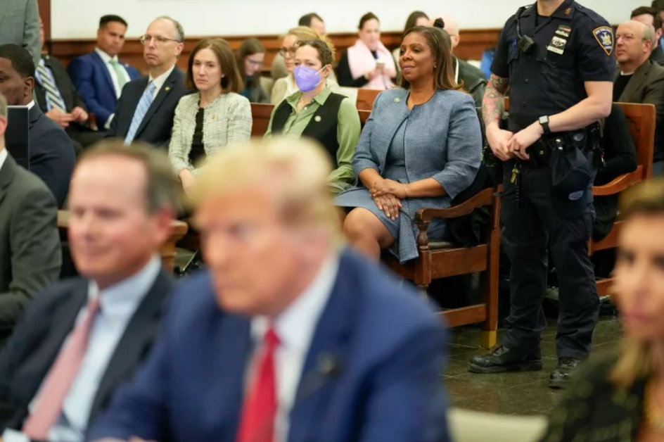 A courtroom  is filled with people seated with both men and woman facing the front. President Trump is scene in the forefront but is out of focus sitting along two people on each side of him. 