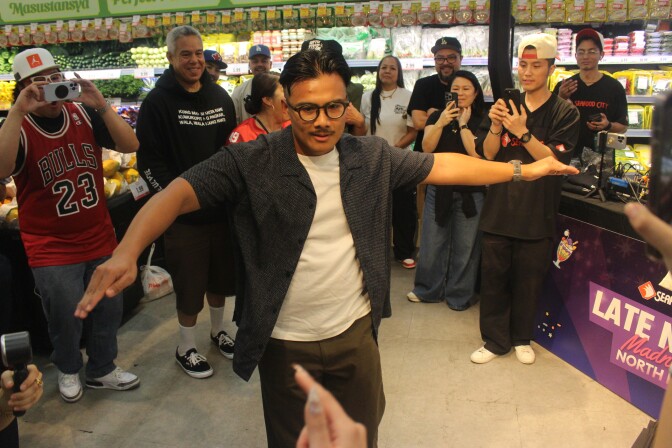 A person in a Seafood City shirt dances with arms outstretched inside a Seafood City supermarket as a crowd watches and cheers during a dance battle.