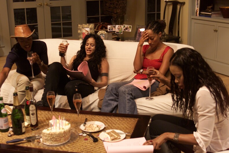 Four Black women are gathered around a coffee table with a birthday cake with candles.