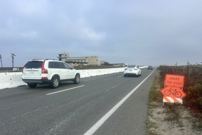 Cars drive on a coastal highway near a 'Share The Road' bicycle sign posted along the shoulder.