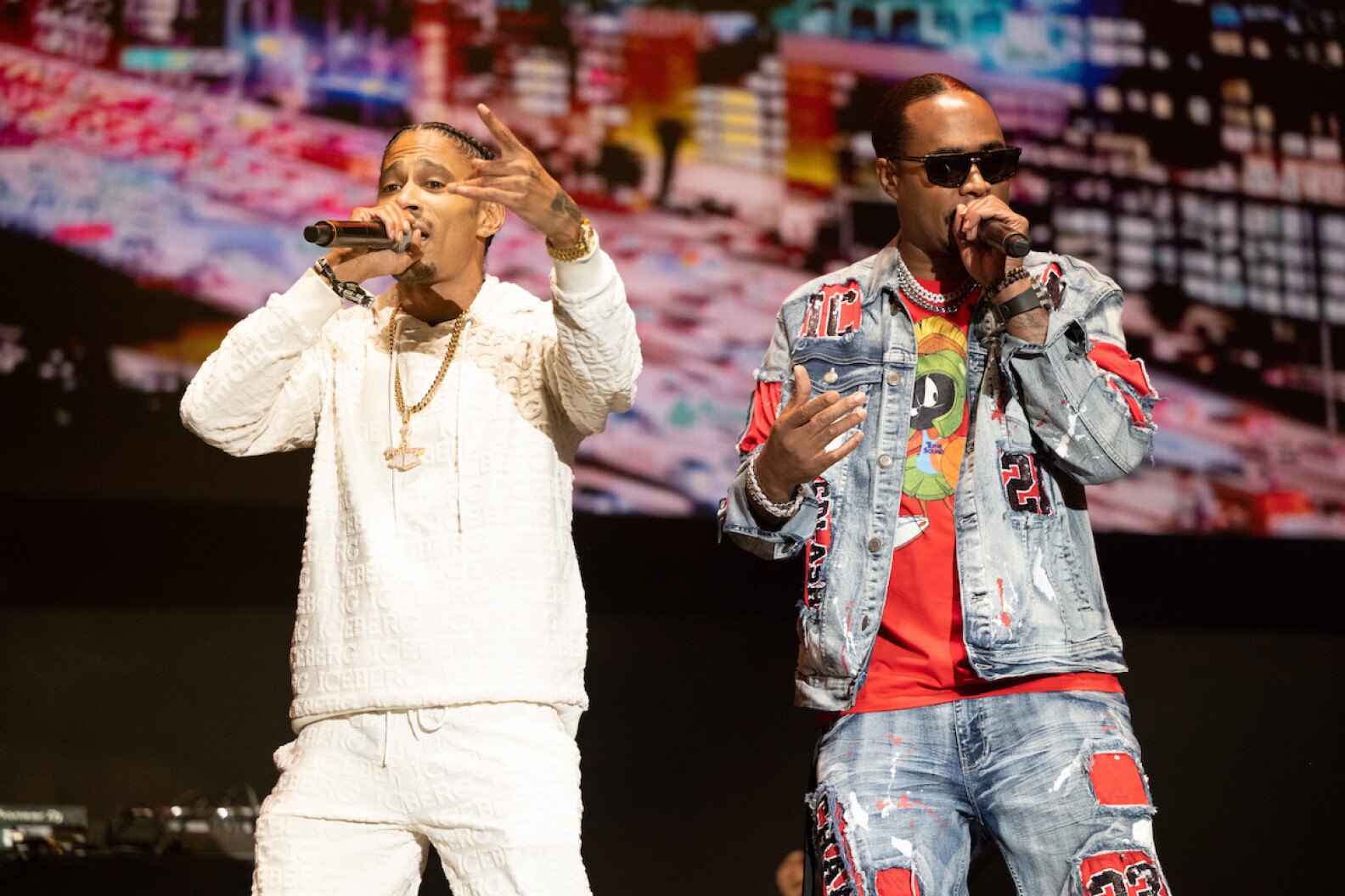 Rapper Layzie Bone, a man wearing all white with a gold chain, performs next to rapper Flesh-N-Bone, a man wearing light denim and orange and black sunglasses.