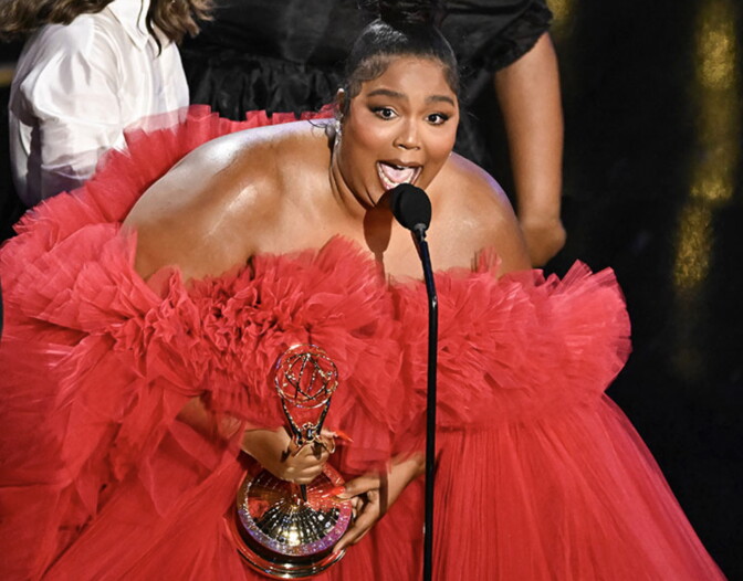 The singer Lizzo in a red dress speaking into a microphone after winning the Emmy for best competitive reality show.