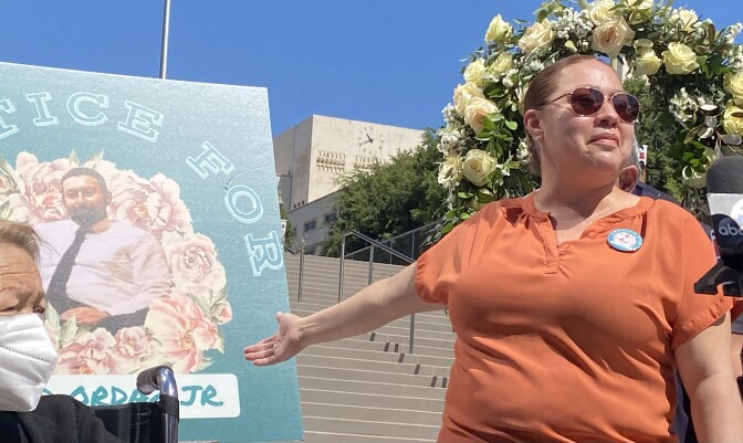 David Ordaz, Jr's sister, Hilda Pedroza, stands in front of flight of stairs outside a downtown LA courthouse at a press conference. She is wearing an orange shirt with a large button with a depiction of Ordaz and is pointing to a painting of the same depiction of Ordaz, who was killed by Los Angeles Sheriff's Deputies on March 14, 2021.