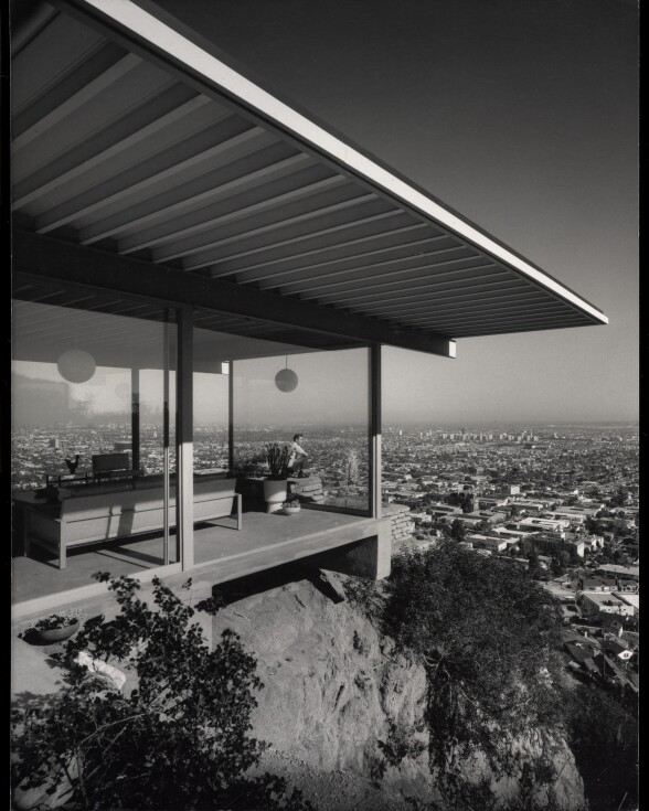 A black and white photo of a modernist house taken from the outside. It's built on top of a steep hillside, overlooking the city below.