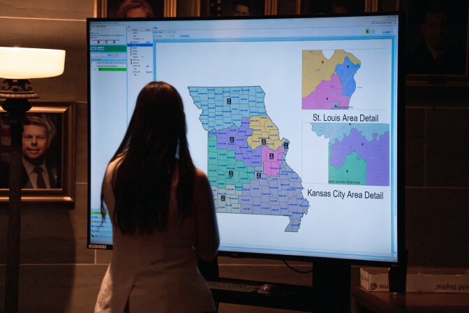 A woman with long dark hair, wearing a beige blazer, is pictured from behind. She is standing in front of a large television monitor that shows three multi colored maps.