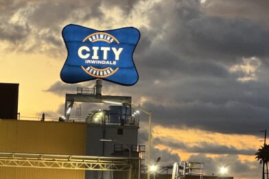 A big blue sign that say "City Brewing & Beverage Irwindale" is seen at sunset. Lights illuminate the sign. Large clouds can be seen behind it. Palm trees and power lines are seen in the right hand corner.