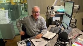 A man with light-tone skin and a button-down shirt and glass rests his arms on a desk in a radio studio where he has papers, multiple screens and a mic with a KPCC 89.3 flag