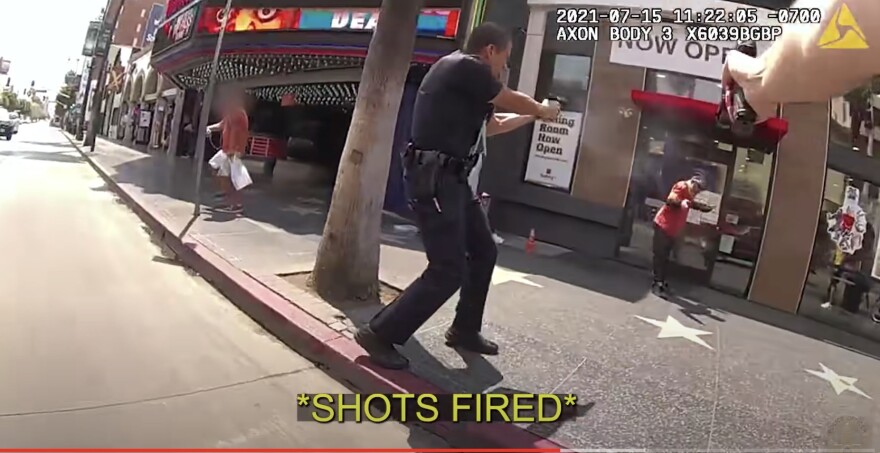 A screen shot from the officer body cam footage of the fatal shooting of Matthew Sova in 2021 shows officers pointing their weapons at Sova, who wears a red shirt and dark pants and is standing in front of a business in Hollywood. 