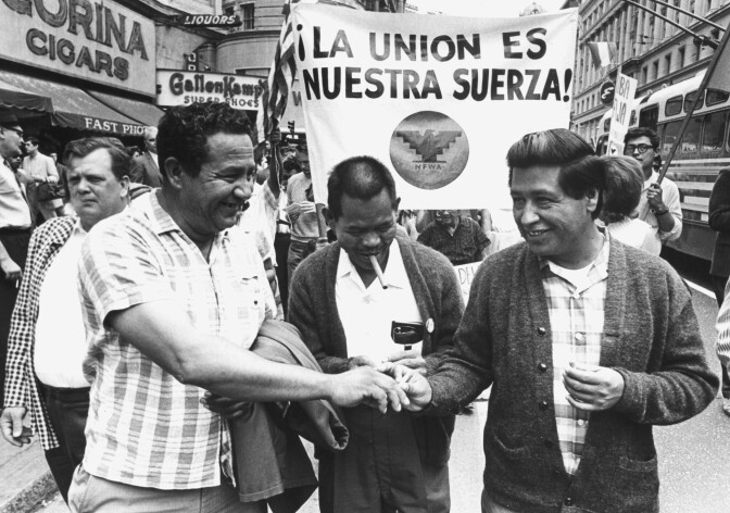 Black and white photo of three men walking in front of a group of protesters. The man in the middle is looking down and a cigarette is in his mouth. The men on either side of him are reaching across him, shaking hands.