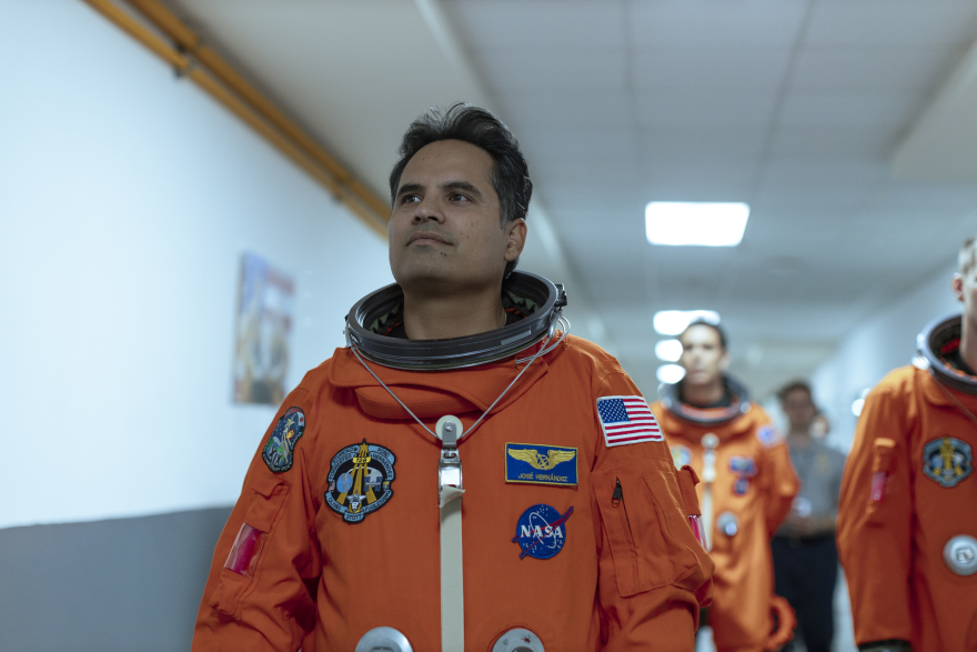 Michael Pena walks down a brightly lit hallway wearing an orange astronaut suit. His chin is slightly raised, a faint smile across his lips, as he looks forward filled with self pride. A trail of other astronauts in the same suits follow behind him.