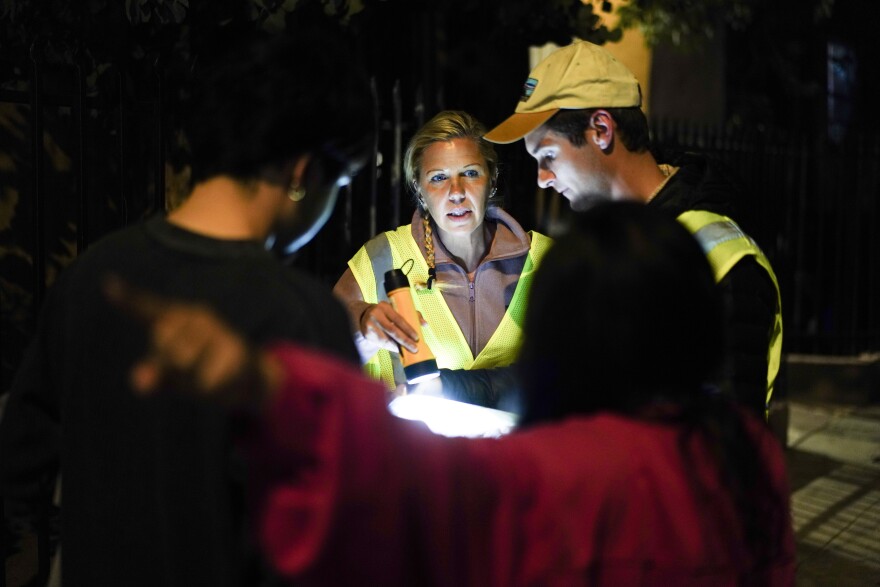 A woman points down the street, showing two people in reflective vests where people have been living on the street.