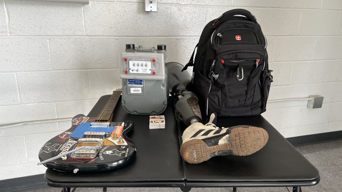 A small black table is seen in a room, there is a white brick wall behind the table and a gray linoleum floor under it. On the table an electric guitar, and electric meter, a stack of baseball cards, a prosthetic leg with an Adidas shoe attached, and a black backpack are depicted.