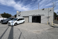 A tan concrete building with the words "Metro Lost & Found" above the entrance is seen. Two cars one gray truck and one white van are seen in the front of the building. A blue sky and various powerlines can be seen in the background.