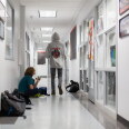 Two students in a bright school hallway: one sits on the floor against the wall eating a snack beside backpacks and a lunch bag, while another in a gray hooded sweatshirt walks away from the camera.