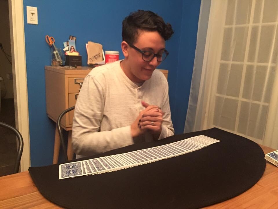 A person with medium-light skin tone and short haircut, wearing glasses and a white top, sits in front of a deck of cards laid out face down on a black felt cloth