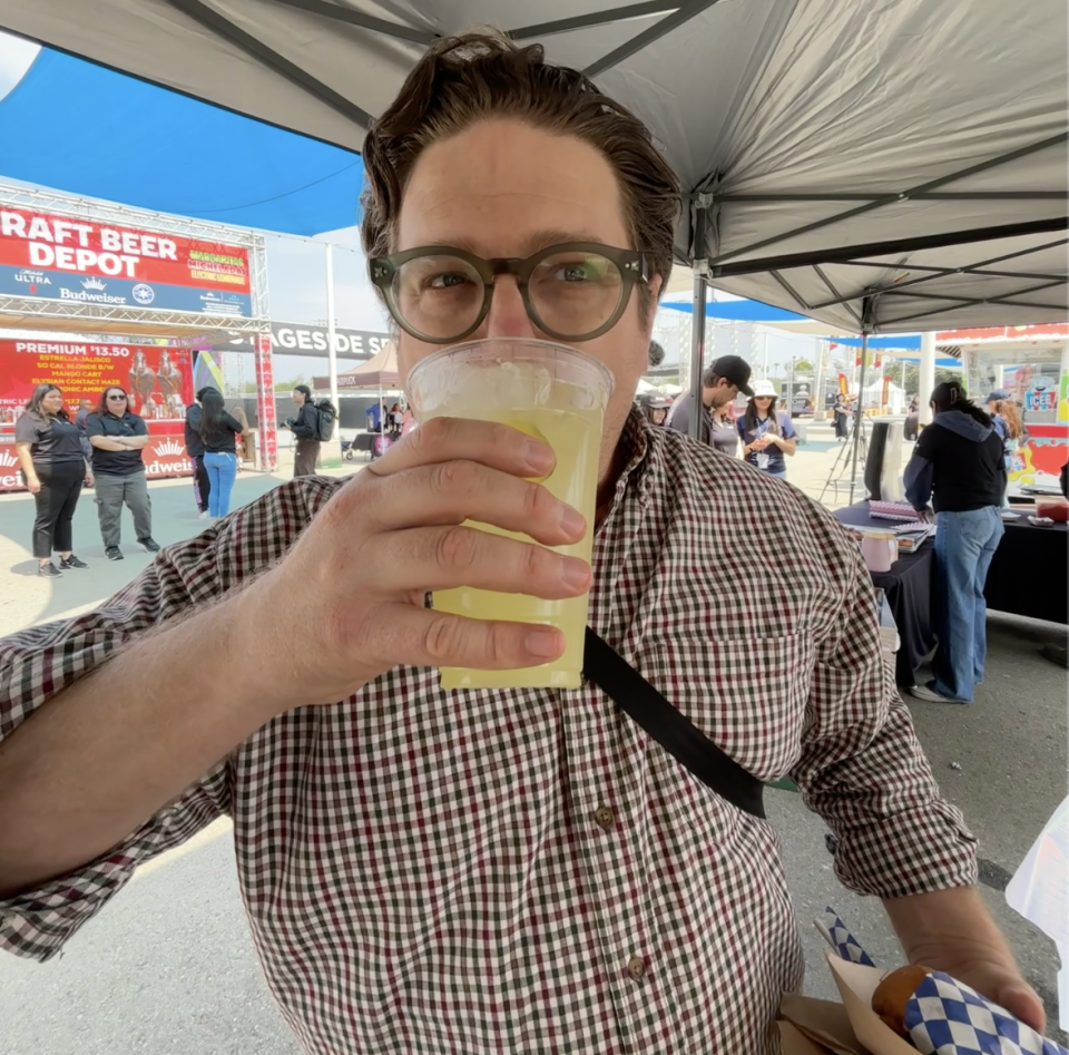 A man with light skin and glasses and brown hair wearing a checkered shirt holding a large clear plastic cup of lemonade up to his lips about to take a sip. 