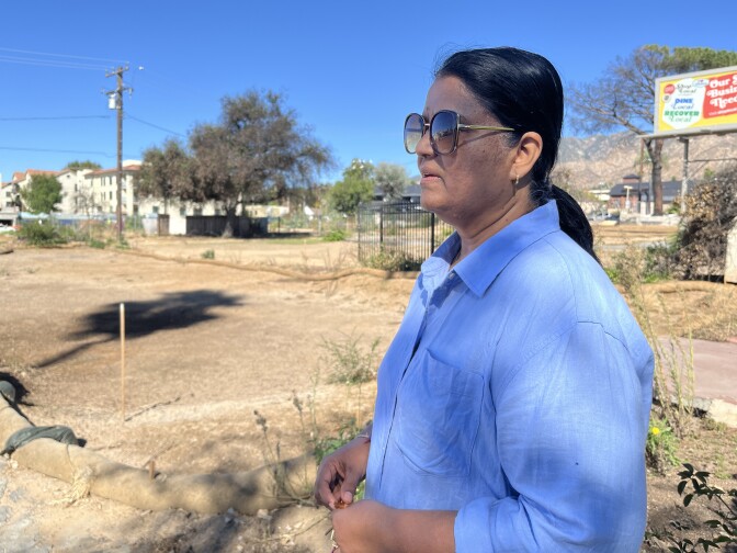 A woman with medium dark skin tone and blue shirt looks to the distance in front of an empty dirt lot.  
