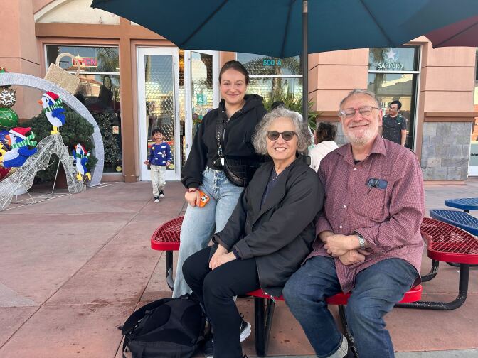 A young light skinned woman wearing blue jeans and a black jacket poses and smiles with an older light skinned woman in a black jacket and an older light skinned man in a red long-sleeve button down shirt. They are sitting at a picnic table in front of a restaurant that has Christmas decorations in the corner. 