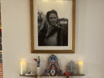 A framed photo of a white woman with long hair is on a wall above a shelf with candles, flowers and other icons