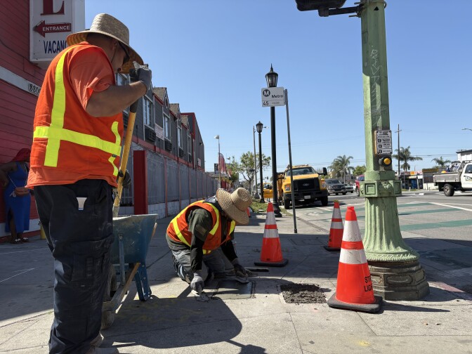There are two men in high-visibility vests and wide-brimmed hats on a road. One is standing next to a blue wheelbarrow with a shovel. The other man is on his knees using a tool to spread wet concrete over box where street lighting wires are stored. 