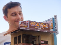 A small version of Al & Bea's Mexican Food in the foreground. It's a brown and tan-colored building with an awning that overlooks a front entrance with metal bars. A small open sign is in the window. On the right side is a wall that has a sign for the business and a Dollar Plus store. The building is being held up by the artist, who is standing in the background.