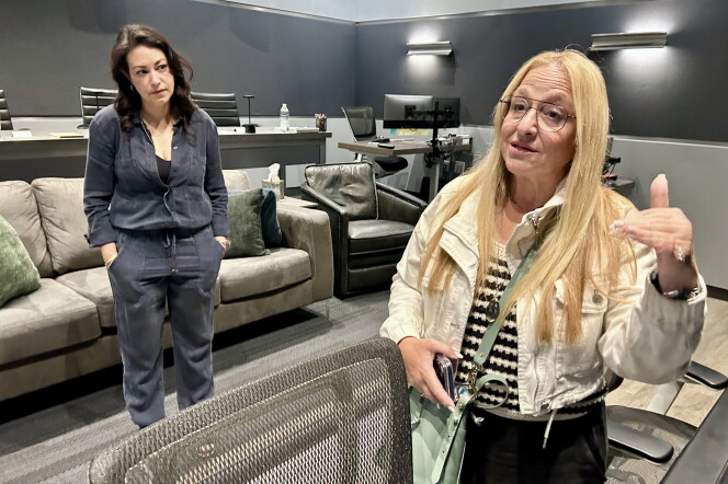 Two female presenting persons stand inside a room painted gray. Several couches are behind them.