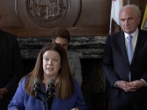 City Attorney Hydee Feldstein Soto, a woman with light skin tone, straight brown hair, wearing a blue blazer stands at a podium and speaks into a microphone. Behind her is Mark Adams, a man with light skin tone, white hair, wearing a navy blue blazer, white shirt, and light blue tie. He stands in front of two flags. 