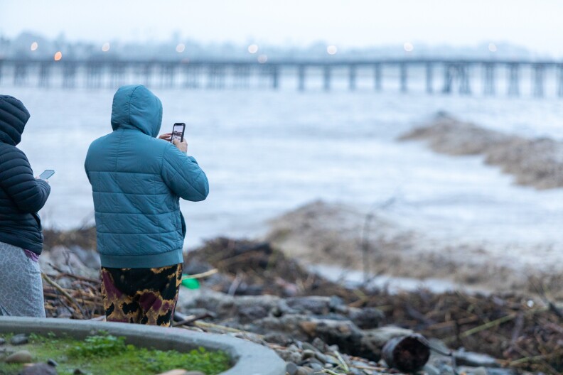 Two men one wearing a blue jacket and the other a black jacket hold their phones as they look out towards the stormy and debris filled ocean water. A pier can be seen out of focus in the background.