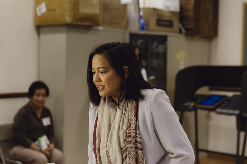 A woman with medium-light skin tone and short dark hair wearing a lavender blazer and a beige and maroon scarf leans towards something out of frame looking downwards.