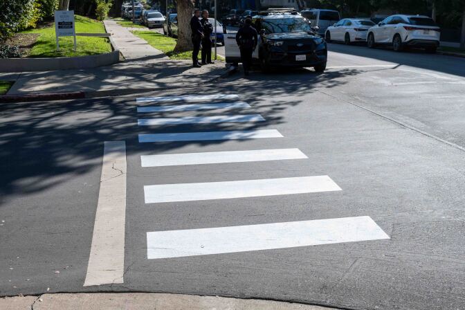 White crosswalk markings are shown on a road on a sunny day. There is a police car at the intersection behind the crosswalk. 