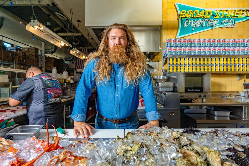 A portrait of a man with long, flowing golden red hair and beard. He is wearing a blue shirt and standing in front of a large display of seafood, covered in ice. Behind him a prep chef cuts onions.