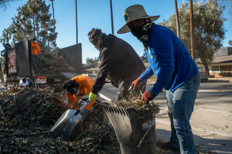 Three people wearing long sleeve shirts and masks are shoveling and picking up debris on a street. Another person in an orange long sleeve shirt wearing a mask stands on a large truck bed filled with debris.
