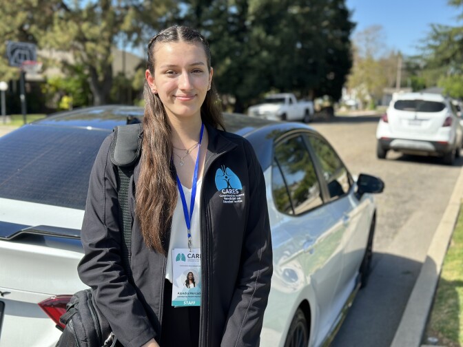 A young woman with light skin tone wears a jacket with an icon of lungs on her left side. She has long brown hair and is smiling at the camera.