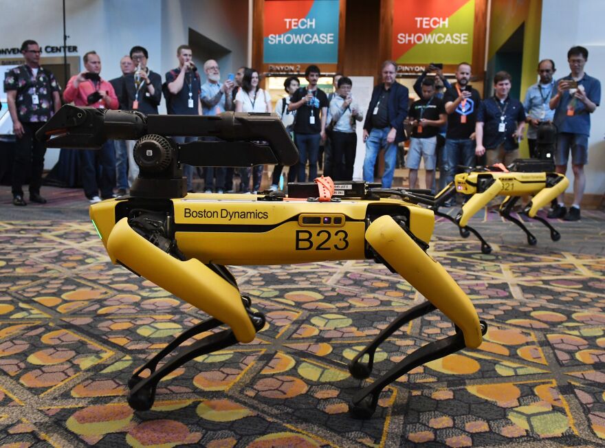 A pair of bright yellow four-legged robots are marked "Boston Dynamics" in a room with heavy patterned carpet as people look on.