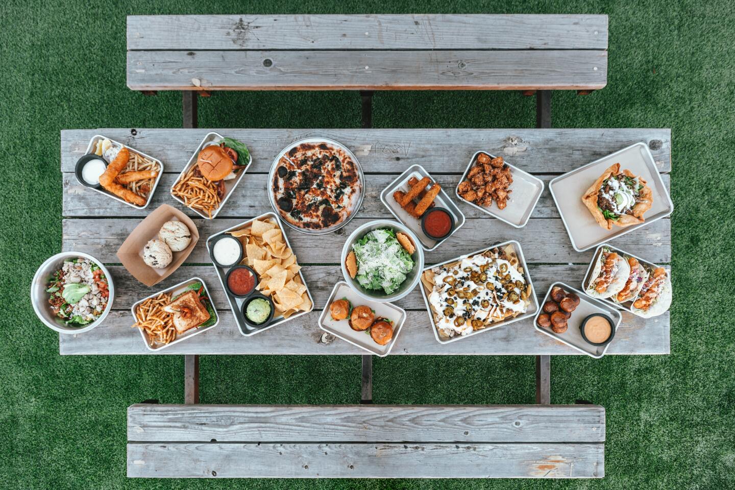 An overhead shot of a picnic table loaded with food: nachos, tacos, a taco salad, fried mozzarella sticks, burgers, a sandwich