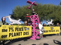 People hold large yellow banners reading "the 1% flies, our planet dies" and "ban private jets." Two people sit on top of a pink wooden tower with a pink plane at the top that has black lettering reading "1%"